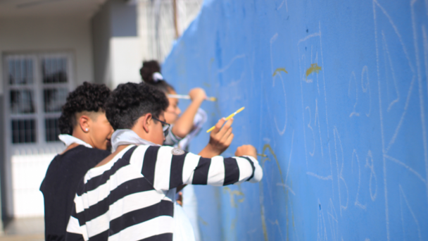 Esta &eacute; uma descri&ccedil;&atilde;o da imagem para fins de acessibilidade:

Descri&ccedil;&atilde;o da Imagem
Uma fotografia colorida em plano m&eacute;dio e &acirc;ngulo lateral captura tr&ecirc;s jovens participando de uma atividade de pintura art&iacute;stica em um muro escolar, parte do projeto Artista na Escola.

A&ccedil;&atilde;o Principal: Os jovens est&atilde;o alinhados ao longo de um muro azul vibrante, utilizando pinc&eacute;is finos de cabo amarelo para preencher ou contornar desenhos e n&uacute;meros previamente esbo&ccedil;ados em branco.

Os Jovens:

Em primeiro plano, um jovem de cabelos escuros e cacheados veste uma camiseta de mangas longas com listras horizontais pretas e brancas. Ele est&aacute; focado na pintura &agrave; sua frente.

Atr&aacute;s dele, um segundo jovem de perfil tamb&eacute;m trabalha no muro.

Ao fundo, uma terceira estudante com o cabelo preso em um coque alto &eacute; vista segurando um pincel.

O Muro: A superf&iacute;cie &eacute; de um tom azul claro intenso. Sobre ela, h&aacute; marca&ccedil;&otilde;es em giz ou tinta branca que lembram um calend&aacute;rio ou grade num&eacute;rica, com n&uacute;meros como "5", "24/31", "28" e "29" vis&iacute;veis.

O Ambiente:

A cena ocorre ao ar livre sob luz natural clara.

O fundo est&aacute; levemente desfocado, mostrando parte da estrutura cinza da escola e uma porta ou port&atilde;o de metal branco com grades.

A profundidade de campo &eacute; reduzida, mantendo o foco principal no jovem com a camiseta listrada e em sua a&ccedil;&atilde;o de pintar.
