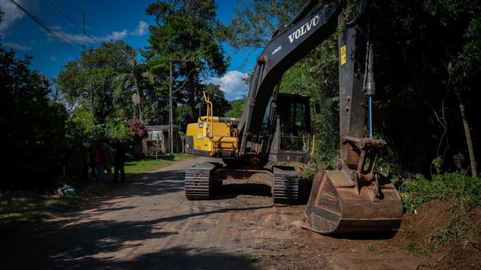 Foto mostra retroescavadeira numa estrada de ch&atilde;o, cercada de &aacute;rvores.