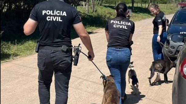 Tr&ecirc;s agentes da Pol&iacute;cia Civil caminham por uma estrada de terra, cada um conduzindo um cachorro com coleira.
