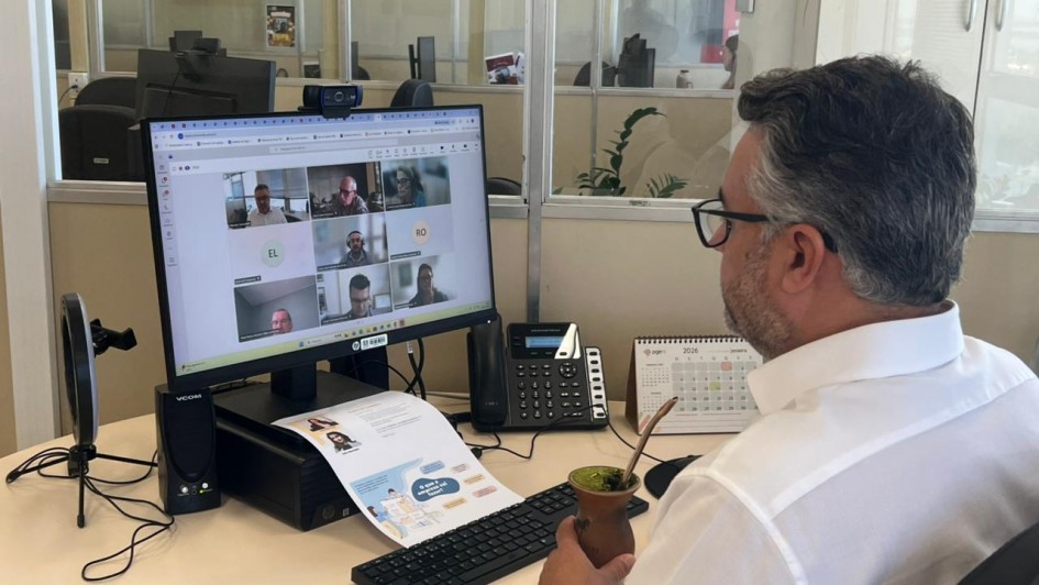 Esta &eacute; uma descri&ccedil;&atilde;o da imagem para fins de acessibilidade:

Descri&ccedil;&atilde;o da Imagem
Uma fotografia colorida em plano m&eacute;dio, capturada de um &acirc;ngulo lateral e por tr&aacute;s (vis&atilde;o sobre o ombro), mostra um profissional participando de uma reuni&atilde;o virtual em um ambiente de escrit&oacute;rio.

O Participante: Um homem de cabelos grisalhos e &oacute;culos de grau, vestindo uma camisa social branca, est&aacute; sentado &agrave; sua mesa de trabalho. Ele segura com a m&atilde;o direita uma cuia de chimarr&atilde;o com boma met&aacute;lica, caracter&iacute;stica da cultura ga&uacute;cha.

O Monitor: &Agrave; frente dele, um monitor de computador exibe uma videochamada em andamento com uma grade de oito participantes (alguns com v&iacute;deo ligado e outros apenas com iniciais como "EL" e "RO").

A Mesa de Trabalho: Sobre a mesa clara, al&eacute;m do monitor e do teclado preto, encontram-se:

Um telefone IP preto.

Um calend&aacute;rio de mesa aberto no m&ecirc;s de janeiro de 2026.

Um documento impresso com ilustra&ccedil;&otilde;es e textos explicativos.

Um pequeno ring light (iluminador circular) montado em um trip&eacute; &agrave; esquerda do monitor.

O Ambiente: O escrit&oacute;rio &eacute; moderno, com divis&oacute;rias de vidro que permitem ver outras esta&ccedil;&otilde;es de trabalho e computadores ao fundo, sugerindo um ambiente de gest&atilde;o p&uacute;blica ou corporativa.