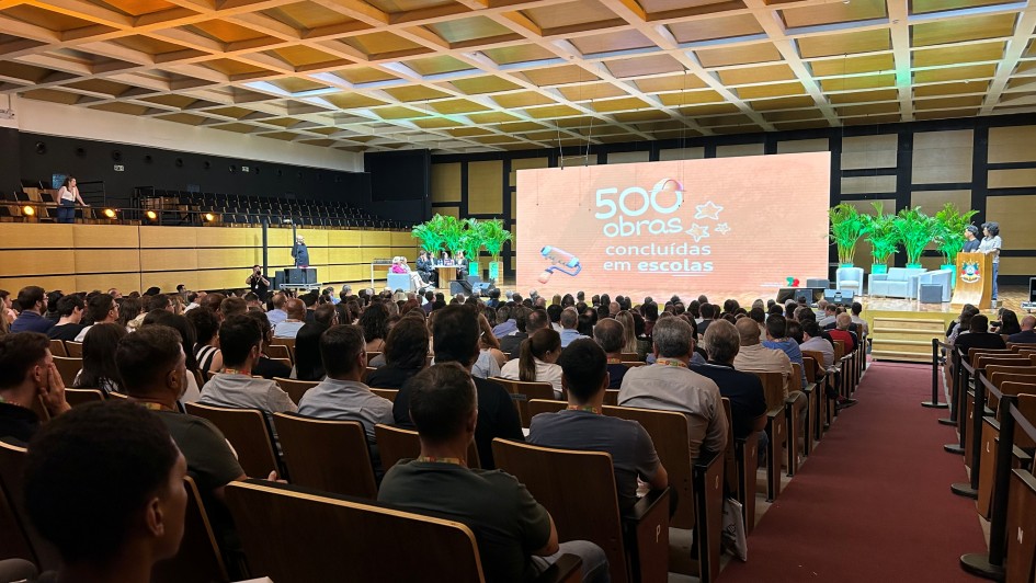 A imagem mostra um audit&oacute;rio lotado. No palco, h&aacute; um tel&atilde;o onde se l&ecirc; "500 obras conclu&iacute;das em escolas". 