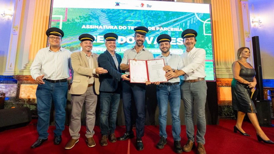 Um grupo de seis homens posa para a foto segurando um documento assinado. Todos sorriem e vestem quepes de maquinista. Ao fundo, h&aacute; um tel&atilde;o onde se l&ecirc; "Assinatura de termo de parceria Trem dos Vales". 