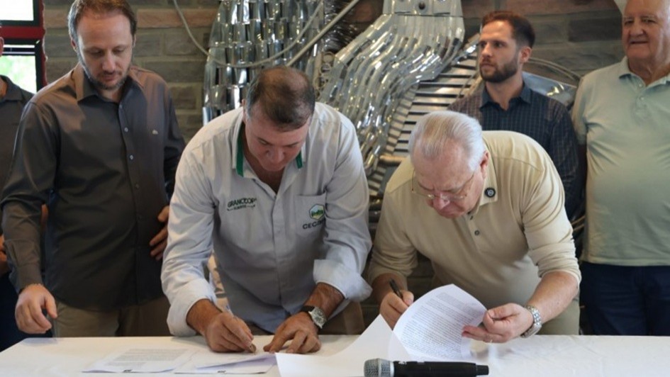 Dois homens est&atilde;o debru&ccedil;ados sobre uma mesa assinando documentos. Ao fundo, aparecem outras pessoas que acompanham o ato. 