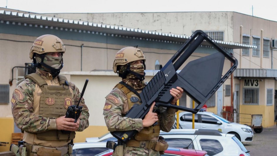 Esta &eacute; uma descri&ccedil;&atilde;o da imagem para fins de acessibilidade:

Descri&ccedil;&atilde;o Geral: Dois agentes da Pol&iacute;cia Penal em trajes t&aacute;ticos de camuflagem est&atilde;o posicionados ao ar livre, um deles segurando um dispositivo tecnol&oacute;gico de grande porte.

Detalhes da Imagem:

Os Agentes: Em primeiro plano, dois agentes vestem fardamento camuflado em tons de bege, marrom e verde. Eles utilizam coletes t&aacute;ticos marrons, cintos de guarni&ccedil;&atilde;o com acess&oacute;rios e capacetes bege. Nos capacetes, h&aacute; um adesivo com a sigla "GAES". Ambos usam balaclavas camufladas que cobrem todo o rosto, deixando apenas os olhos vis&iacute;veis. No bra&ccedil;o, &eacute; vis&iacute;vel o bras&atilde;o da "POL&Iacute;CIA PENAL".

Equipamento Especializado: O agente &agrave; direita segura um equipamento eletr&ocirc;nico preto, grande e de design futurista, com formato retangular vazado, semelhante a uma arma de bloqueio de drones. O agente &agrave; esquerda segura um r&aacute;dio comunicador preto.

O Ambiente: O cen&aacute;rio &eacute; um p&aacute;tio externo, possivelmente em uma unidade prisional. Ao fundo, h&aacute; carros estacionados (incluindo um branco e um vermelho) e uma edifica&ccedil;&atilde;o de cor bege com janelas pequenas e a inscri&ccedil;&atilde;o "MV3" na parede. O c&eacute;u est&aacute; encoberto e a luz &eacute; difusa.