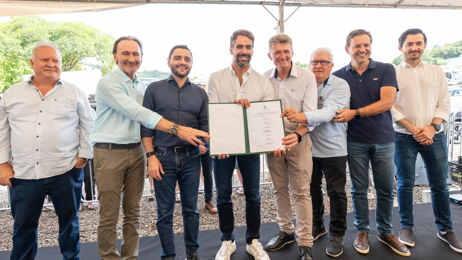 Esta &eacute; uma descri&ccedil;&atilde;o da imagem para fins de acessibilidade:

Descri&ccedil;&atilde;o Geral: O governador Eduardo Leite e um grupo de sete homens posam para uma foto oficial em um palco, segurando juntos um documento assinado que autoriza o in&iacute;cio de obras em uma rodovia.

Detalhes da Imagem:

As Pessoas: No centro da imagem, o governador Eduardo Leite (homem jovem, de barba aparada, vestindo camisa social branca e cal&ccedil;a jeans escura) segura uma pasta verde aberta. Ao seu redor, outros sete homens de diferentes idades, vestindo camisas sociais (em tons de azul, branco e cinza) e cal&ccedil;as casuais, sorriem para a c&acirc;mera. Alguns deles tamb&eacute;m seguram as bordas da pasta ou apoiam as m&atilde;os uns nos outros, demonstrando coopera&ccedil;&atilde;o.

O Documento: A pasta verde exibe um papel branco com textos e diversas assinaturas manuscritas, simbolizando o ato oficial de autoriza&ccedil;&atilde;o das obras na ERS-332.

O Ambiente: O grupo est&aacute; posicionado sobre um tablado coberto com um tecido preto. O evento ocorre ao ar livre, sob a prote&ccedil;&atilde;o de uma grande tenda de lona branca.

O Plano de Fundo: Atr&aacute;s do palco, v&ecirc;-se uma &aacute;rea externa com ch&atilde;o de brita, &aacute;rvores verdes ao fundo e a luz clara de um dia ensolarado. A composi&ccedil;&atilde;o da foto &eacute; frontal e foca na uni&atilde;o do grupo em torno do documento.