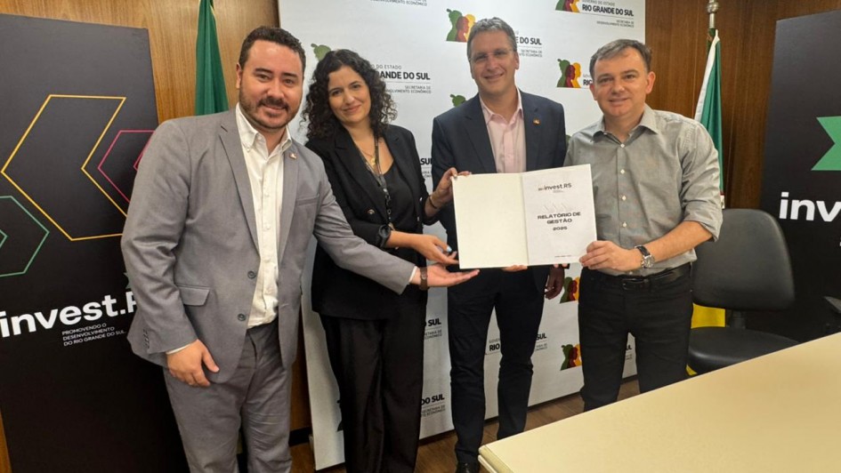 Esta &eacute; uma descri&ccedil;&atilde;o da imagem para fins de acessibilidade:

Descri&ccedil;&atilde;o Geral: Quatro representantes da ag&ecirc;ncia Invest RS e da Secretaria de Desenvolvimento Econ&ocirc;mico posam para uma foto oficial em um escrit&oacute;rio, segurando juntos um relat&oacute;rio de gest&atilde;o de 2025.

Detalhes da Imagem:

As Pessoas: O grupo &eacute; formado por tr&ecirc;s homens e uma mulher, todos sorridentes e em trajes profissionais. Da esquerda para a direita: um homem de terno cinza e barba; uma mulher de blazer preto e cabelos cacheados; um homem de palet&oacute; azul-marinho; e um homem de camisa social cinza.

O Documento: No centro da imagem, eles seguram coletivamente uma pasta branca. Na capa, est&atilde;o impressos o logotipo "invest.RS" e o t&iacute;tulo "RELAT&Oacute;RIO DE GEST&Atilde;O 2025".

O Plano de Fundo: Atr&aacute;s do grupo, h&aacute; um painel branco com v&aacute;rias reprodu&ccedil;&otilde;es do logotipo do Governo do Estado do Rio Grande do Sul e da Secretaria de Desenvolvimento Econ&ocirc;mico. Tamb&eacute;m s&atilde;o vis&iacute;veis bandeiras oficiais (incluindo a do Rio Grande do Sul) e banners pretos com a identidade visual da Invest RS nas laterais.

Composi&ccedil;&atilde;o: A cena registra um ato formal de presta&ccedil;&atilde;o de contas em um ambiente administrativo com paredes revestidas de madeira, transmitindo uma atmosfera de coopera&ccedil;&atilde;o e transpar&ecirc;ncia.