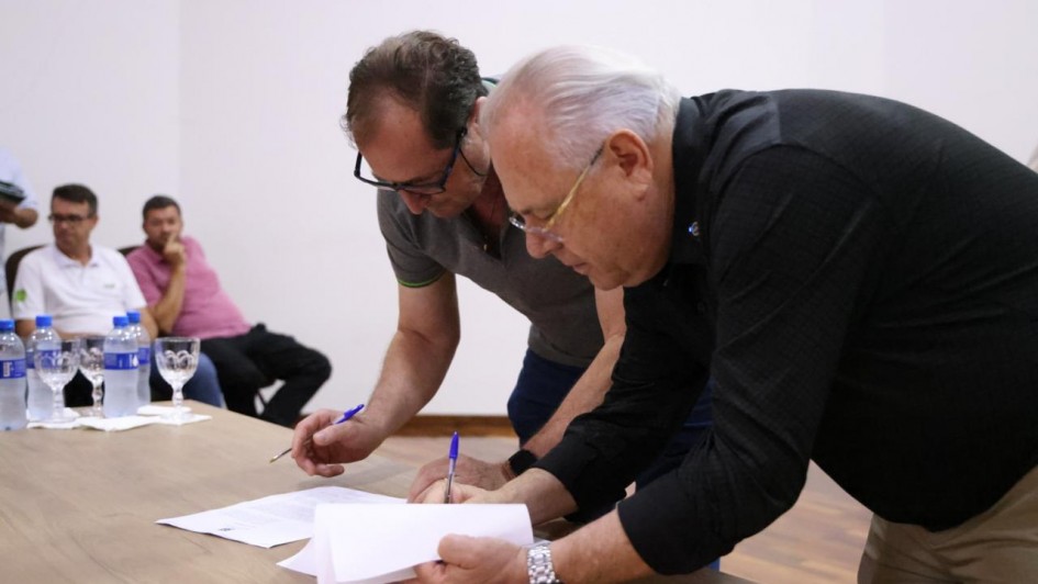 Dois homens est&atilde;o curvados sobre uma mesa assinando documentos, enquanto outras pessoas observam ao fundo.