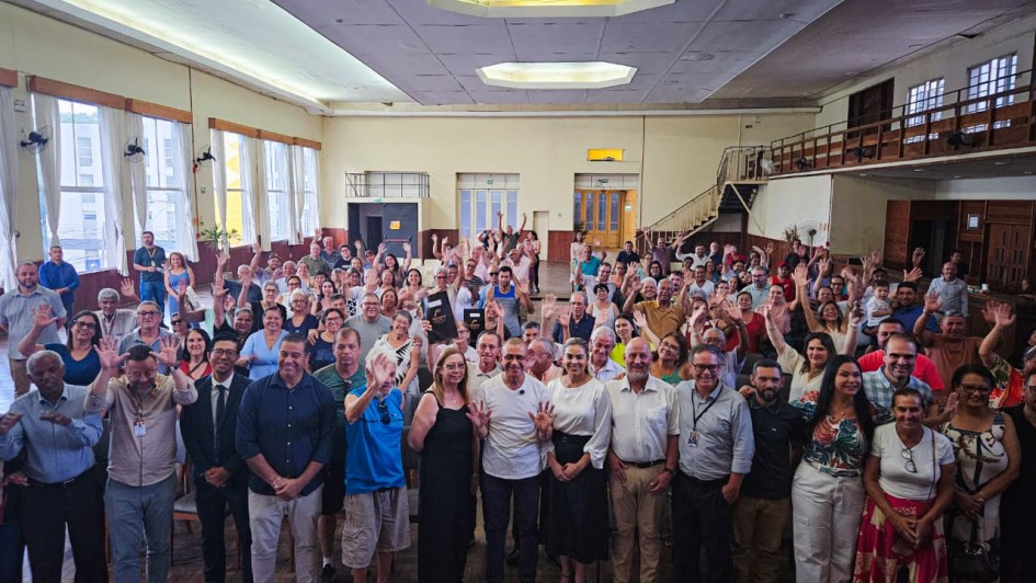 Um grande grupo de pessoas est&aacute; reunido em um sal&atilde;o amplo e iluminado, posando para a foto e sorrindo, com v&aacute;rias delas acenando para a c&acirc;mera.
