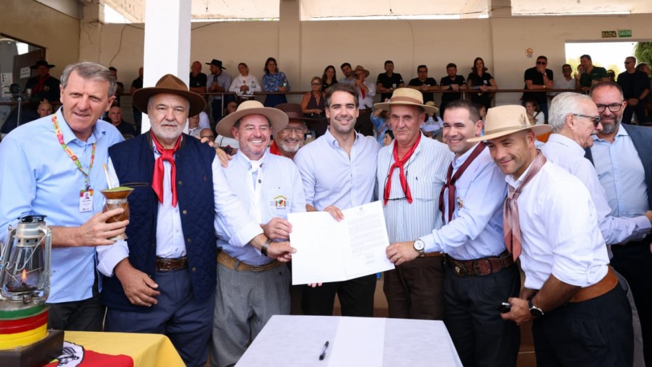 Um grupo de homens sorridentes, muitos usando trajes t&iacute;picos ga&uacute;chos com len&ccedil;os e chap&eacute;us, posa segurando um documento aberto em um evento com p&uacute;blico ao fundo.