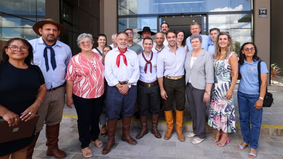 Um grupo de homens e mulheres posa sorrindo em frente a um pr&eacute;dio moderno; alguns usam trajes t&iacute;picos ga&uacute;chos com bombachas, len&ccedil;os e botas.