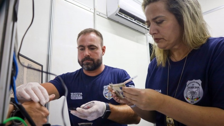 Esta imagem captura o trabalho t&eacute;cnico e minucioso de emiss&atilde;o de documentos de identidade no Rio Grande do Sul. Aqui est&aacute; uma descri&ccedil;&atilde;o detalhada:

Composi&ccedil;&atilde;o da Imagem
A foto mostra dois profissionais do Instituto-Geral de Per&iacute;cias (IGP) em um ambiente de escrit&oacute;rio ou posto de atendimento, focados na coleta de dados biom&eacute;tricos e confer&ecirc;ncia de documentos.

Elementos Principais
Os Profissionais:

&Agrave; esquerda, um homem de barba curta e camiseta azul-marinho com o logotipo da "Per&iacute;cia Criminal RS" est&aacute; concentrado em coletar as impress&otilde;es digitais de uma pessoa (cuja m&atilde;o aparece parcialmente no canto inferior esquerdo). Ele usa luvas brancas de procedimento para garantir a higiene e a qualidade da coleta.

&Agrave; direita, uma mulher tamb&eacute;m veste o uniforme azul-marinho do IGP. Ela segura e analisa atentamente um documento em papel, possivelmente uma certid&atilde;o ou formul&aacute;rio necess&aacute;rio para o processo de identifica&ccedil;&atilde;o.

O Ambiente:

O cen&aacute;rio &eacute; funcional, com divis&oacute;rias brancas t&iacute;picas de postos de atendimento p&uacute;blico.

No canto superior, v&ecirc;-se um aparelho de ar-condicionado instalado na parede.

Sobre a mesa, h&aacute; equipamentos t&eacute;cnicos, incluindo o que parece ser um scanner de digitais e outros materiais de escrit&oacute;rio.

Detalhes de Identifica&ccedil;&atilde;o:

Ambos os servidores utilizam uniformes com o bras&atilde;o oficial do Instituto-Geral de Per&iacute;cias do RS, refor&ccedil;ando o car&aacute;ter oficial e institucional da cena.

A imagem transmite uma sensa&ccedil;&atilde;o de seriedade, precis&atilde;o e servi&ccedil;o p&uacute;blico, ilustrando o esfor&ccedil;o log&iacute;stico mencionado no t&iacute;tulo do arquivo para atender &agrave; alta demanda por novas carteiras de identidade.