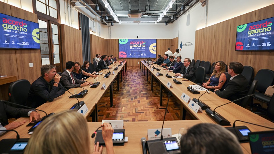 Esta imagem registra uma reuni&atilde;o t&eacute;cnica e institucional organizada pelo Governo do Estado do Rio Grande do Sul. Abaixo, uma descri&ccedil;&atilde;o detalhada da cena:

Cen&aacute;rio e Composi&ccedil;&atilde;o
A foto apresenta uma sala de confer&ecirc;ncias ampla e moderna, com um design que mistura elementos cl&aacute;ssicos e contempor&acirc;neos.

Mobili&aacute;rio: Uma grande mesa de madeira em formato de "U" ocupa o centro da sala, permitindo que todos os participantes se vejam de frente.

Piso e Paredes: O ch&atilde;o &eacute; revestido com parquet de madeira em padr&atilde;o espinha de peixe. As paredes possuem pain&eacute;is de madeira clara, conferindo um aspecto s&oacute;brio e profissional ao ambiente.

Identidade Visual: Em destaque, h&aacute; tr&ecirc;s telas de TV (duas laterais e uma ao fundo) exibindo a marca "acordo ga&uacute;cho - edital n&ordm; 02/25 | ICMS". O logotipo utiliza cores vibrantes como vermelho, amarelo e verde, e inclui o &iacute;cone de um aperto de m&atilde;os, simbolizando concilia&ccedil;&atilde;o.

Os Participantes
Disposi&ccedil;&atilde;o: Cerca de 15 a 20 pessoas, vestidas predominantemente em trajes formais (palet&oacute;s e camisas sociais), est&atilde;o sentadas ao redor da mesa.

Intera&ccedil;&atilde;o: Os participantes parecem engajados em uma apresenta&ccedil;&atilde;o ou discuss&atilde;o. Alguns utilizam microfones de mesa individuais, e h&aacute; pastas, pap&eacute;is e copos d'&aacute;gua distribu&iacute;dos &agrave; frente de cada um.

Representatividade: Nota-se a presen&ccedil;a de homens e mulheres de diferentes faixas et&aacute;rias, caracterizando uma equipe t&eacute;cnica de governo (Secretaria da Fazenda e Procuradoria-Geral do Estado, conforme sugerido pelos logotipos nas telas).

Detalhes T&eacute;cnicos e Ambiente
Ilumina&ccedil;&atilde;o: O teto possui uma estrutura de trilhos industriais com lumin&aacute;rias modernas, garantindo uma ilumina&ccedil;&atilde;o clara e uniforme.

Tecnologia: Al&eacute;m das telas de exibi&ccedil;&atilde;o, a mesa est&aacute; equipada com sistemas de &aacute;udio individuais para confer&ecirc;ncia, refor&ccedil;ando a natureza oficial do evento.

A imagem comunica um ambiente de transpar&ecirc;ncia, di&aacute;logo e gest&atilde;o p&uacute;blica, focado na resolu&ccedil;&atilde;o estrat&eacute;gica de quest&otilde;es tribut&aacute;rias.