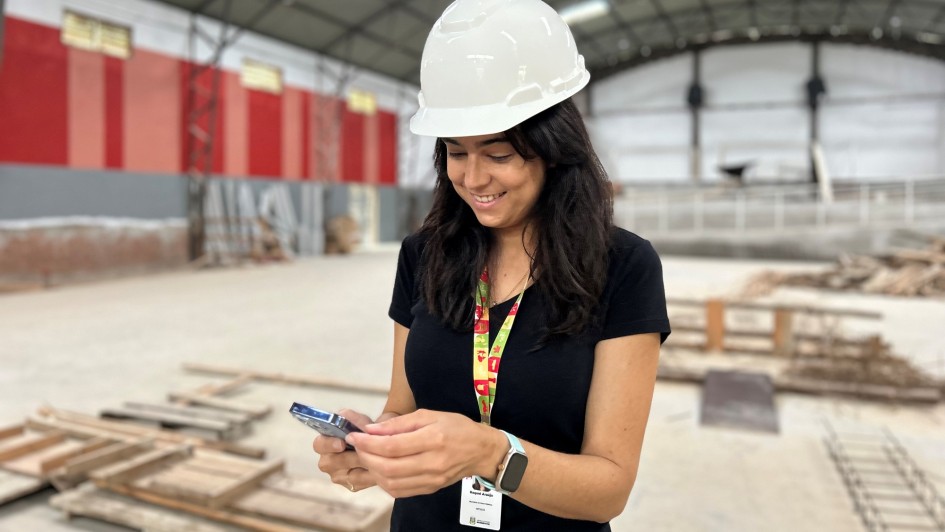 Foto mostra arquiteta com capacete branco e usando celular em um canteiro de obras. 