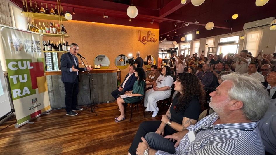 A imagem mostra um homem em p&eacute; em frente a um microfone, falando para uma plateia sentada em um ambiente de bar. No canto esquerdo, h&aacute; um banner com a marca dos 400 anos das Miss&otilde;es e da Secretaria da Cultura do RS. 