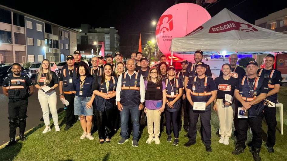 Esta &eacute; uma descri&ccedil;&atilde;o detalhada da imagem para fins de acessibilidade:

Descri&ccedil;&atilde;o Geral
A fotografia registra um grupo de aproximadamente 20 pessoas reunidas &agrave; noite em uma &aacute;rea gramada ao lado de uma rua urbana. O grupo celebra os 15 anos da Opera&ccedil;&atilde;o Balada Segura em uma a&ccedil;&atilde;o comemorativa no litoral.

Elementos em Destaque
O Grupo: Homens e mulheres de diversas idades est&atilde;o posicionados em p&eacute;, lado a lado, formando tr&ecirc;s fileiras para a foto oficial.

Vestimentas: * A maioria veste coletes refletivos azul-marinho com faixas laranjas sobre camisetas escuras, uniforme padr&atilde;o dos agentes de tr&acirc;nsito.

Alguns integrantes usam camisetas pretas com o logotipo circular vermelho e branco da "Balada Segura".

&Agrave; esquerda, um homem veste um uniforme t&aacute;tico preto completo.

Express&otilde;es: Os participantes olham para a c&acirc;mera com express&otilde;es serenas e profissionais, transmitindo um sentimento de uni&atilde;o e dever cumprido.

Cen&aacute;rio e Objetos
Lado Direito: H&aacute; uma tenda branca de lona com o logotipo "BALADA SEGURA" em vermelho. Atr&aacute;s da tenda, destaca-se um grande bal&atilde;o infl&aacute;vel rosa iluminado, onde se l&ecirc; "Prefeitura Municipal" e o s&iacute;mbolo da campanha (um boneco estilizado segurando um volante).

Lado Esquerdo: Uma rua asf&aacute;ltica com carros estacionados e pr&eacute;dios residenciais iluminados ao fundo.

Ilumina&ccedil;&atilde;o: A cena &eacute; iluminada por luzes artificiais urbanas e pelo brilho interno do bal&atilde;o infl&aacute;vel, criando sombras n&iacute;tidas no gramado em primeiro plano.