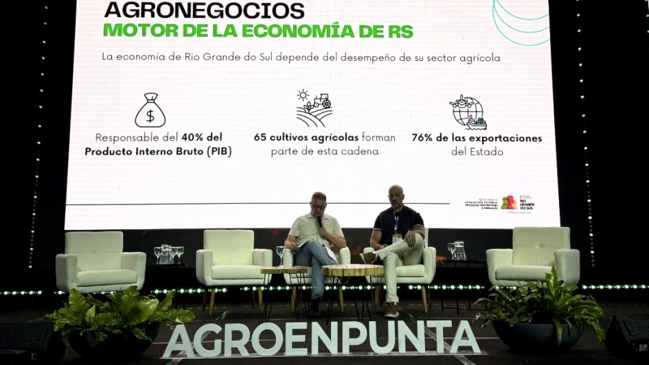 Aqui est&aacute; a descri&ccedil;&atilde;o detalhada da imagem para fins de acessibilidade:

A imagem registra uma apresenta&ccedil;&atilde;o ou painel de discuss&atilde;o em um palco com ilumina&ccedil;&atilde;o c&ecirc;nica. No centro, uma grande tela de proje&ccedil;&atilde;o exibe dados sobre a import&acirc;ncia do agroneg&oacute;cio.

O Palco e os Participantes: Dois homens est&atilde;o sentados em poltronas brancas confort&aacute;veis, posicionadas no centro do palco. Eles parecem estar em uma conversa ou apresenta&ccedil;&atilde;o formal. No total, h&aacute; quatro poltronas brancas e pequenas mesas de apoio com copos e jarras de &aacute;gua. &Agrave; frente do palco, no ch&atilde;o, l&ecirc;-se em letras grandes e claras a palavra "AGROENPUNTA", ladeada por dois vasos com folhagens verdes densas.

A Tela de Proje&ccedil;&atilde;o: O slide em destaque possui fundo branco e texto em espanhol.

T&iacute;tulo: "AGRONEGOCIOS MOTOR DE LA ECONOM&Iacute;A DE RS" (Agroneg&oacute;cios Motor da Economia do RS).

Subt&iacute;tulo: "La econom&iacute;a de Rio Grande do Sul depende del desempe&ntilde;o de su sector agr&iacute;cola".

Infogr&aacute;fico com tr&ecirc;s pontos principais:

&Iacute;cone de saco de dinheiro: "Responsable del 40% del Producto Interno Bruto (PIB)".

&Iacute;cone de trator e campo: "65 cultivos agr&iacute;colas forman parte de esta cadena".

&Iacute;cone de navio cargueiro e globo: "76% de las exportaciones del Estado".

Ilumina&ccedil;&atilde;o e Ambiente: O ambiente ao redor da tela &eacute; escuro, com fileiras de luzes LED verdes decorando tanto a parte superior quanto a base do painel de proje&ccedil;&atilde;o. No canto inferior direito da tela, aparecem os logotipos do Governo do Estado do Rio Grande do Sul e da Secretaria da Agricultura, Pecu&aacute;ria, Produ&ccedil;&atilde;o Sustent&aacute;vel e Irriga&ccedil;&atilde;o.