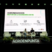 Aqui est&aacute; a descri&ccedil;&atilde;o detalhada da imagem para fins de acessibilidade:

A imagem registra uma apresenta&ccedil;&atilde;o ou painel de discuss&atilde;o em um palco com ilumina&ccedil;&atilde;o c&ecirc;nica. No centro, uma grande tela de proje&ccedil;&atilde;o exibe dados sobre a import&acirc;ncia do agroneg&oacute;cio.

O Palco e os Participantes: Dois homens est&atilde;o sentados em poltronas brancas confort&aacute;veis, posicionadas no centro do palco. Eles parecem estar em uma conversa ou apresenta&ccedil;&atilde;o formal. No total, h&aacute; quatro poltronas brancas e pequenas mesas de apoio com copos e jarras de &aacute;gua. &Agrave; frente do palco, no ch&atilde;o, l&ecirc;-se em letras grandes e claras a palavra "AGROENPUNTA", ladeada por dois vasos com folhagens verdes densas.

A Tela de Proje&ccedil;&atilde;o: O slide em destaque possui fundo branco e texto em espanhol.

T&iacute;tulo: "AGRONEGOCIOS MOTOR DE LA ECONOM&Iacute;A DE RS" (Agroneg&oacute;cios Motor da Economia do RS).

Subt&iacute;tulo: "La econom&iacute;a de Rio Grande do Sul depende del desempe&ntilde;o de su sector agr&iacute;cola".

Infogr&aacute;fico com tr&ecirc;s pontos principais:

&Iacute;cone de saco de dinheiro: "Responsable del 40% del Producto Interno Bruto (PIB)".

&Iacute;cone de trator e campo: "65 cultivos agr&iacute;colas forman parte de esta cadena".

&Iacute;cone de navio cargueiro e globo: "76% de las exportaciones del Estado".

Ilumina&ccedil;&atilde;o e Ambiente: O ambiente ao redor da tela &eacute; escuro, com fileiras de luzes LED verdes decorando tanto a parte superior quanto a base do painel de proje&ccedil;&atilde;o. No canto inferior direito da tela, aparecem os logotipos do Governo do Estado do Rio Grande do Sul e da Secretaria da Agricultura, Pecu&aacute;ria, Produ&ccedil;&atilde;o Sustent&aacute;vel e Irriga&ccedil;&atilde;o.