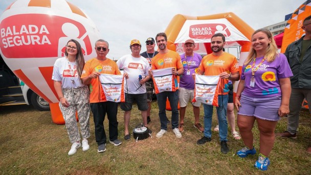 Aqui est&aacute; a descri&ccedil;&atilde;o detalhada da imagem para fins de acessibilidade:

A imagem registra um evento institucional realizado em um gramado ao ar livre sob c&eacute;u nublado, com a presen&ccedil;a de autoridades do governo do Rio Grande do Sul.

Figuras Centrais e A&ccedil;&atilde;o: No centro da composi&ccedil;&atilde;o, um grupo de oito pessoas posa para a foto. O governador Eduardo Leite e o vice-governador Gabriel Souza, acompanhados de um terceiro homem, seguram &agrave; frente do corpo mochilas do tipo saco de cor branca. Nas mochilas, l&ecirc;-se em destaque a frase: "Na estrada, o salva-vidas &eacute; voc&ecirc;.".

Vestimentas e Identidade Visual: * Eduardo Leite e Gabriel Souza vestem camisetas laranjas com o logotipo da campanha "Ver&atilde;o Total".

Outros integrantes do grupo usam camisetas roxas, tamb&eacute;m com a marca do "Ver&atilde;o Total", ou camisetas brancas com o logo da "Balada Segura".

Elementos de Fundo: * &Agrave; esquerda, h&aacute; um grande infl&aacute;vel em formato esf&eacute;rico (semelhante a um capacete ou bal&atilde;o), nas cores laranja e branco, exibindo a marca "BALADA SEGURA" e o &iacute;cone de uma pessoa com cinto de seguran&ccedil;a.

Ao fundo, uma estrutura infl&aacute;vel laranja em formato de tenda tamb&eacute;m exibe o logotipo da "Balada Segura".

Uma parte de um ve&iacute;culo branco &eacute; vis&iacute;vel na extremidade esquerda da foto.
