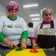 Duas mulheres vestindo aventais e toucas est&atilde;o picando tomates e temperos em uma bancada. Uma veste camiseta identificada com a marca do projeto Emancipa Fam&iacute;lia Ga&uacute;cha. 