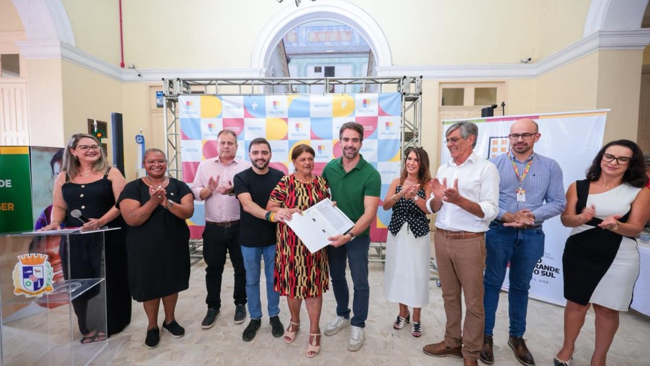 A imagem registra um momento de celebra&ccedil;&atilde;o e reconhecimento esportivo durante um evento oficial do governo estadual no litoral.

A&ccedil;&atilde;o Central: O governador Eduardo Leite (&agrave; esquerda) e o vice-governador Gabriel Souza (ao centro) posam para uma foto ao lado de um jovem atleta. Eles seguram, em conjunto, uma prancha de surfe branca com detalhes em cinza e azul, e o atleta segura um certificado ou diploma de reconhecimento.

As Pessoas:

Eduardo Leite: Veste uma camisa polo azul-marinho e sorri para a c&acirc;mera.

Gabriel Souza: Veste a camiseta laranja da "Opera&ccedil;&atilde;o Ver&atilde;o Total", mantendo a identidade visual vista em fotos anteriores.

O Atleta: Um jovem loiro, vestindo uma camiseta preta com estampas de marcas de surfe (como a marca "Mormaii"), segurando o certificado com orgulho.

Ao fundo: Outras pessoas acompanham o ato, incluindo uma mulher de &oacute;culos e blazer claro que tamb&eacute;m apareceu em registros de inaugura&ccedil;&otilde;es de sa&uacute;de anteriores.

O Cen&aacute;rio: O evento ocorre sob uma estrutura de lona branca (tenda). Ao fundo, h&aacute; um painel azul com logotipos institucionais, onde se l&ecirc; parte da palavra "GOVERNO" e o bras&atilde;o do Estado do Rio Grande do Sul. O ch&atilde;o &eacute; de areia, confirmando que a atividade acontece na orla da praia.

Elementos Visuais: A prancha de surfe tem adesivos de patrocinadores e marcas esportivas. A ilumina&ccedil;&atilde;o &eacute; natural de um dia nublado, sem sombras marcadas.