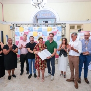 A imagem registra um momento de celebra&ccedil;&atilde;o e reconhecimento esportivo durante um evento oficial do governo estadual no litoral.

A&ccedil;&atilde;o Central: O governador Eduardo Leite (&agrave; esquerda) e o vice-governador Gabriel Souza (ao centro) posam para uma foto ao lado de um jovem atleta. Eles seguram, em conjunto, uma prancha de surfe branca com detalhes em cinza e azul, e o atleta segura um certificado ou diploma de reconhecimento.

As Pessoas:

Eduardo Leite: Veste uma camisa polo azul-marinho e sorri para a c&acirc;mera.

Gabriel Souza: Veste a camiseta laranja da "Opera&ccedil;&atilde;o Ver&atilde;o Total", mantendo a identidade visual vista em fotos anteriores.

O Atleta: Um jovem loiro, vestindo uma camiseta preta com estampas de marcas de surfe (como a marca "Mormaii"), segurando o certificado com orgulho.

Ao fundo: Outras pessoas acompanham o ato, incluindo uma mulher de &oacute;culos e blazer claro que tamb&eacute;m apareceu em registros de inaugura&ccedil;&otilde;es de sa&uacute;de anteriores.

O Cen&aacute;rio: O evento ocorre sob uma estrutura de lona branca (tenda). Ao fundo, h&aacute; um painel azul com logotipos institucionais, onde se l&ecirc; parte da palavra "GOVERNO" e o bras&atilde;o do Estado do Rio Grande do Sul. O ch&atilde;o &eacute; de areia, confirmando que a atividade acontece na orla da praia.

Elementos Visuais: A prancha de surfe tem adesivos de patrocinadores e marcas esportivas. A ilumina&ccedil;&atilde;o &eacute; natural de um dia nublado, sem sombras marcadas.