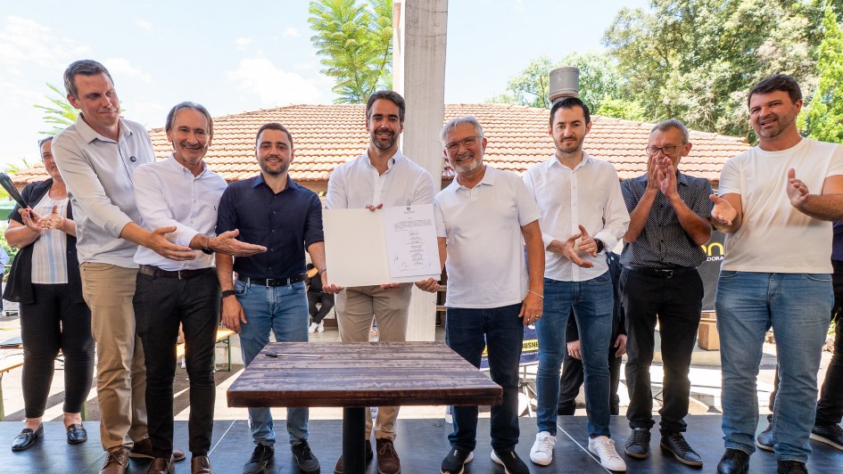 A fotografia registra o momento da assinatura da ordem de in&iacute;cio para obras de recupera&ccedil;&atilde;o em rodovias do Vale do Ca&iacute;.

Elementos Principais:
Centro da Imagem: O Governador Eduardo Leite est&aacute; posicionado ao centro, vestindo uma camisa social branca de mangas longas e cal&ccedil;a bege. Ele segura e exibe uma pasta branca aberta, mostrando o documento assinado para a c&acirc;mera.

Grupo de Acompanhamento: Ao lado do governador, est&atilde;o outros oito homens e uma mulher (parcialmente vis&iacute;vel na extremidade esquerda). Eles vestem trajes casuais e sociais, predominantemente camisas brancas ou escuras e cal&ccedil;as jeans. Alguns sorriem, enquanto um homem &agrave; direita faz um gesto de aplauso.

Cen&aacute;rio: O grupo est&aacute; sobre um tablado ou palco de madeira escura ao ar livre. &Agrave; frente deles, h&aacute; uma pequena mesa quadrada de madeira.

Ambiente e Fundo:
Constru&ccedil;&atilde;o: Ao fundo, destaca-se uma edifica&ccedil;&atilde;o com telhado de telhas de barro vermelho.

Natureza: A cena &eacute; emoldurada por &aacute;rvores verdes e folhagens sob um c&eacute;u azul claro, indicando um dia ensolarado.