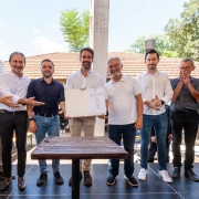 A fotografia registra o momento da assinatura da ordem de in&iacute;cio para obras de recupera&ccedil;&atilde;o em rodovias do Vale do Ca&iacute;.

Elementos Principais:
Centro da Imagem: O Governador Eduardo Leite est&aacute; posicionado ao centro, vestindo uma camisa social branca de mangas longas e cal&ccedil;a bege. Ele segura e exibe uma pasta branca aberta, mostrando o documento assinado para a c&acirc;mera.

Grupo de Acompanhamento: Ao lado do governador, est&atilde;o outros oito homens e uma mulher (parcialmente vis&iacute;vel na extremidade esquerda). Eles vestem trajes casuais e sociais, predominantemente camisas brancas ou escuras e cal&ccedil;as jeans. Alguns sorriem, enquanto um homem &agrave; direita faz um gesto de aplauso.

Cen&aacute;rio: O grupo est&aacute; sobre um tablado ou palco de madeira escura ao ar livre. &Agrave; frente deles, h&aacute; uma pequena mesa quadrada de madeira.

Ambiente e Fundo:
Constru&ccedil;&atilde;o: Ao fundo, destaca-se uma edifica&ccedil;&atilde;o com telhado de telhas de barro vermelho.

Natureza: A cena &eacute; emoldurada por &aacute;rvores verdes e folhagens sob um c&eacute;u azul claro, indicando um dia ensolarado.