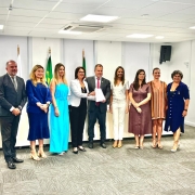 Esta &eacute; uma descri&ccedil;&atilde;o detalhada da imagem para fins de acessibilidade:

Vis&atilde;o Geral
A imagem registra um momento formal em um ambiente de escrit&oacute;rio ou sala de reuni&otilde;es. Nove pessoas &mdash; tr&ecirc;s homens e seis mulheres &mdash; est&atilde;o posicionadas lado a lado para uma foto oficial. O clima &eacute; profissional e solene, com todos sorrindo levemente para a c&acirc;mera.

Composi&ccedil;&atilde;o e Elementos Centrais
A&ccedil;&atilde;o Principal: No centro da imagem, uma mulher vestindo um blazer branco e cal&ccedil;as pretas e um homem em um terno escuro com gravata vermelha seguram juntos um documento em papel branco. Este gesto simboliza a assinatura ou entrega do termo mencionado no t&iacute;tulo.

As Pessoas (da esquerda para a direita):

Um homem de terno cinza escuro e gravata.

Uma mulher com um conjunto de vestido e casaco azul vibrante.

Uma mulher com um macac&atilde;o azul-celeste sem mangas.

A mulher de blazer branco (mencionada na a&ccedil;&atilde;o central).

O homem de gravata vermelha (mencionado na a&ccedil;&atilde;o central).

Uma mulher vestindo blusa e cal&ccedil;as brancas.

Uma mulher com um vestido midi marrom.

Uma mulher com um vestido curto de listras verticais em tons de laranja e marrom.

Uma mulher com um vestido azul-marinho abotoado na frente.

Cen&aacute;rio e Ambiente
O Local: Uma sala ampla com piso de carpete cinza em padr&atilde;o quadriculado. Ao fundo, h&aacute; uma mesa branca comprida e tr&ecirc;s mastros com bandeiras (incluindo a bandeira do Brasil e, possivelmente, a do Estado).

Ilumina&ccedil;&atilde;o: A luz &eacute; branca e uniforme, vinda de pain&eacute;is de LED no teto de gesso.

Detalhes Adicionais: &Agrave; direita, h&aacute; uma porta branca e uma placa na parede onde se l&ecirc; parcialmente "DEFENSORIA DO ESTADO".