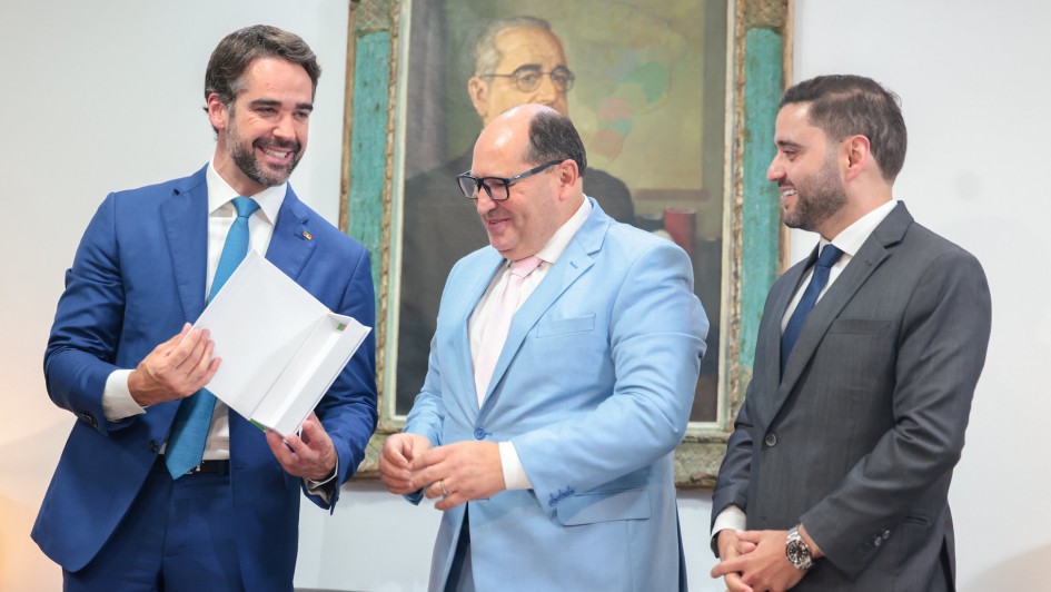 Vis&atilde;o Geral
A imagem captura um momento formal e descontra&iacute;do entre tr&ecirc;s homens em um ambiente institucional. Eles est&atilde;o posicionados &agrave; frente de uma parede branca, onde se destaca um retrato hist&oacute;rico ao fundo.

Pessoas e Elementos Centrais
Homem &agrave; esquerda: O governador Eduardo Leite aparece sorridente, vestindo um terno azul vivo e uma gravata azul-clara. Ele segura com as duas m&atilde;os um livro ou documento de capa branca aberto, apresentando-o aos demais.

Homem ao centro: Veste um terno azul-claro, camisa branca e gravata rosa. Ele usa &oacute;culos de arma&ccedil;&atilde;o escura e olha para baixo, em dire&ccedil;&atilde;o ao documento que est&aacute; sendo exibido.

Homem &agrave; direita: Veste um terno cinza-chumbo e gravata escura. Ele sorri e mant&eacute;m as m&atilde;os cruzadas &agrave; frente do corpo, observando a intera&ccedil;&atilde;o.

Cen&aacute;rio e Composi&ccedil;&atilde;o
Ambiente: O fundo &eacute; uma parede branca simples e bem iluminada. No centro, atr&aacute;s do grupo, h&aacute; uma pintura emoldurada retratando um homem de &oacute;culos (figura hist&oacute;rica).

Detalhes adicionais: No canto esquerdo da imagem, &eacute; poss&iacute;vel ver parte de um microfone, sugerindo que o momento ocorre durante um evento oficial ou coletiva de imprensa.