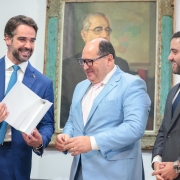 Vis&atilde;o Geral
A imagem captura um momento formal e descontra&iacute;do entre tr&ecirc;s homens em um ambiente institucional. Eles est&atilde;o posicionados &agrave; frente de uma parede branca, onde se destaca um retrato hist&oacute;rico ao fundo.

Pessoas e Elementos Centrais
Homem &agrave; esquerda: O governador Eduardo Leite aparece sorridente, vestindo um terno azul vivo e uma gravata azul-clara. Ele segura com as duas m&atilde;os um livro ou documento de capa branca aberto, apresentando-o aos demais.

Homem ao centro: Veste um terno azul-claro, camisa branca e gravata rosa. Ele usa &oacute;culos de arma&ccedil;&atilde;o escura e olha para baixo, em dire&ccedil;&atilde;o ao documento que est&aacute; sendo exibido.

Homem &agrave; direita: Veste um terno cinza-chumbo e gravata escura. Ele sorri e mant&eacute;m as m&atilde;os cruzadas &agrave; frente do corpo, observando a intera&ccedil;&atilde;o.

Cen&aacute;rio e Composi&ccedil;&atilde;o
Ambiente: O fundo &eacute; uma parede branca simples e bem iluminada. No centro, atr&aacute;s do grupo, h&aacute; uma pintura emoldurada retratando um homem de &oacute;culos (figura hist&oacute;rica).

Detalhes adicionais: No canto esquerdo da imagem, &eacute; poss&iacute;vel ver parte de um microfone, sugerindo que o momento ocorre durante um evento oficial ou coletiva de imprensa.