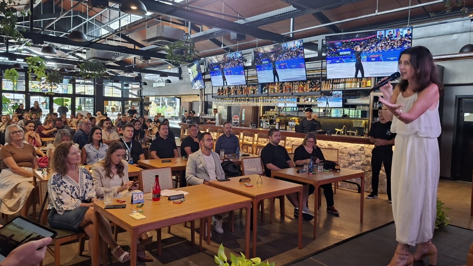Vis&atilde;o Geral
A imagem captura uma palestra ou apresenta&ccedil;&atilde;o realizada em um ambiente informal e moderno, semelhante a um bar ou restaurante. Uma mulher est&aacute; em p&eacute; &agrave; direita, falando para uma plateia sentada em mesas de madeira.

A Oradora e sua A&ccedil;&atilde;o
Posicionamento: No lado direito da imagem, uma mulher de cabelos castanhos ondulados de comprimento m&eacute;dio est&aacute; de p&eacute; sobre um pequeno tablado ou &aacute;rea de destaque.

Vestimenta: Ela veste um vestido longo, branco e fluido, sem mangas.

A&ccedil;&atilde;o: Ela segura um microfone preto com a m&atilde;o direita e faz um gesto aberto com a m&atilde;o esquerda enquanto fala, demonstrando uma postura comunicativa e engajada.

O P&uacute;blico e o Ambiente
A Plateia: Diversas pessoas de idades variadas est&atilde;o sentadas em mesas de madeira clara, distribu&iacute;das pelo sal&atilde;o. Elas est&atilde;o voltadas para a oradora, com express&otilde;es de aten&ccedil;&atilde;o. Algumas pessoas t&ecirc;m copos, pap&eacute;is e celulares sobre as mesas.

O Local: O estabelecimento possui um design industrial e contempor&acirc;neo. O teto &eacute; alto, com vigas met&aacute;licas escuras, lumin&aacute;rias pendentes em formato de c&uacute;pula e plantas decorativas suspensas em cestos.

O Fundo: Ao fundo, h&aacute; um balc&atilde;o de bar iluminado com prateleiras repletas de garrafas de bebidas. Acima do bar, quatro grandes televisores de tela plana est&atilde;o ligados, exibindo o que parecem ser transmiss&otilde;es de competi&ccedil;&otilde;es esportivas.

Ilumina&ccedil;&atilde;o: A luz do ambiente &eacute; quente e suave, criando uma atmosfera acolhedora para o evento.