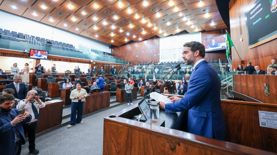 Homem de terno azul fala em um p&uacute;lpito dentro de um grande plen&aacute;rio, com v&aacute;rias pessoas assistindo e tirando fotos.