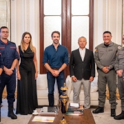 Esta &eacute; uma descri&ccedil;&atilde;o detalhada da imagem para fins de acessibilidade:

Vis&atilde;o Geral
A imagem registra um momento oficial em que oito pessoas &mdash; sete homens e uma mulher &mdash; posam para uma fotografia em um ambiente luxuoso e formal. O grupo est&aacute; organizado em uma linha reta, transmitindo uma atmosfera de coopera&ccedil;&atilde;o institucional.

Pessoas e Vestimentas (da esquerda para a direita)
Bombeiros Militares: Os dois primeiros homens &agrave; esquerda vestem uniformes operacionais azul-marinho com detalhes e identifica&ccedil;&otilde;es em vermelho, t&iacute;picos do Corpo de Bombeiros.

Civil: Uma mulher com cabelos loiros longos veste um vestido preto longo e elegante.

Governador: No centro da imagem, Eduardo Leite veste uma camisa social azul-marinho com as mangas levemente dobradas e cal&ccedil;a jeans escura.

Civil/Autoridade: &Agrave; direita do governador, um homem de cabelos grisalhos veste um blazer preto, camisa branca e cal&ccedil;a cinza-clara.

Brigada Militar: Os dois homens seguintes vestem fardas cinza-oliva da Brigada Militar, com diversos distintivos e ins&iacute;gnias.

T&aacute;tico: Na extremidade direita, um homem veste um uniforme t&aacute;tico camuflado em tons de cinza e preto, equipado com colete operacional.

Cen&aacute;rio e Composi&ccedil;&atilde;o
O Ambiente: O encontro ocorre em uma sala de recep&ccedil;&atilde;o cl&aacute;ssica, possivelmente no Pal&aacute;cio Piratini. O fundo apresenta uma grande porta dupla de madeira com pain&eacute;is de vidro fosco, paredes brancas decoradas com boiseries (molduras de gesso) e espelhos laterais.

Mobili&aacute;rio: Nas laterais, veem-se partes de sof&aacute;s brancos. &Agrave; frente do grupo, h&aacute; uma mesa de centro de madeira escura contendo livros, uma pequena x&iacute;cara de caf&eacute; sobre um pires branco e um vaso decorativo alongado em azul-marinho com detalhes dourados.

Ilumina&ccedil;&atilde;o: A luz &eacute; suave e bem distribu&iacute;da, real&ccedil;ando os detalhes dos uniformes e do ambiente.