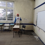 Homem de costas trabalha na parede de uma sala de aula com paredes, piso, pintura e móveis desgastados