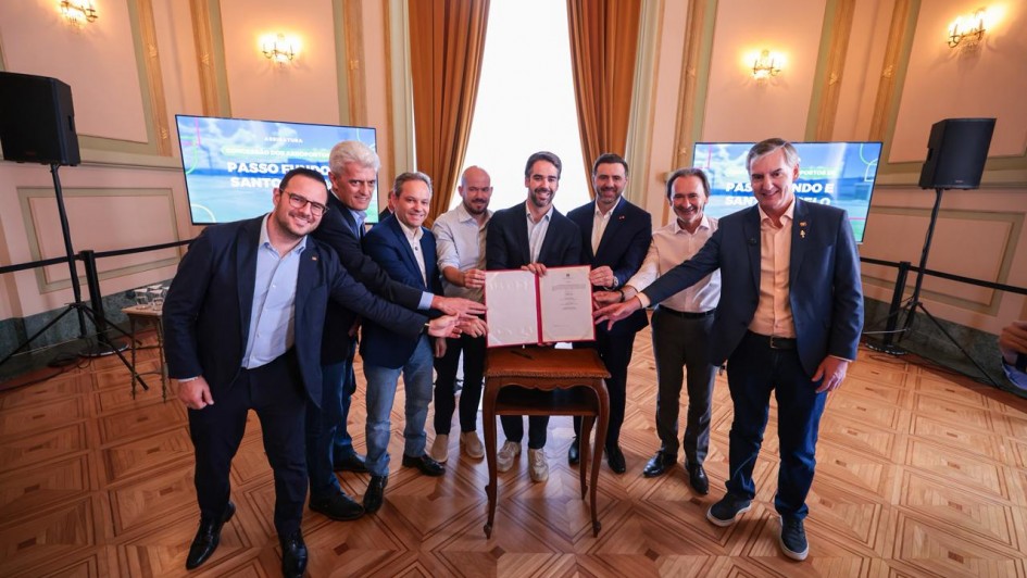 A imagem registra um grupo de pessoas alinhado em frente a uma pequena mesa de madeira posicionada no centro do sal&atilde;o, exibindo um documento aberto com capa vermelha. Todas estendem as m&atilde;os em dire&ccedil;&atilde;o ao documento assinado. 