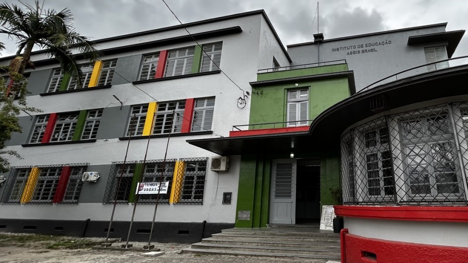 Pr&eacute;dio de tr&ecirc;s andares com v&aacute;rias janelas e pintura nova, com uma grande porta branca na entrada. Acima, letreiro "Instituto de Educa&ccedil;&atilde;o Assis Brasil" 