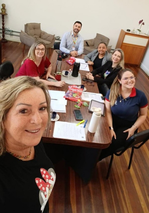 Grupo de cinco pessoas sentadas ao redor de uma mesa com mais uma pessoa em pé, em primeiro plano, como quem tira a foto de todos pelo celular.
