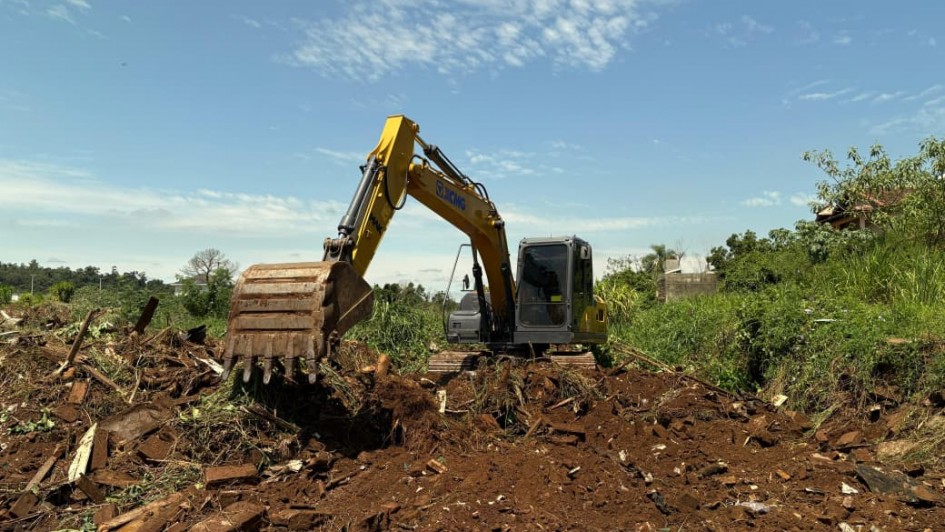 A imagem mostra uma retroescavadeira em a&ccedil;&atilde;o em meio a entulhos e &aacute;reas verdes. 