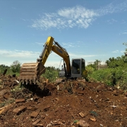 A imagem mostra uma retroescavadeira em a&ccedil;&atilde;o em meio a entulhos e &aacute;reas verdes. 