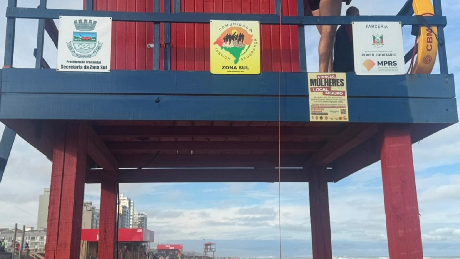 Um posto de salva-vidas vermelho e azul est&aacute; montado na areia da praia, com banhistas caminhando e pr&eacute;dios ao fundo sob um c&eacute;u parcialmente nublado.