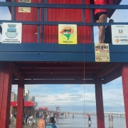Um posto de salva-vidas vermelho e azul est&aacute; montado na areia da praia, com banhistas caminhando e pr&eacute;dios ao fundo sob um c&eacute;u parcialmente nublado.