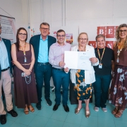 Esta &eacute; uma descri&ccedil;&atilde;o detalhada da imagem para fins de acessibilidade:

Vis&atilde;o Geral
A imagem mostra um grupo de sete pessoas (quatro mulheres e tr&ecirc;s homens) posando para uma foto oficial em um ambiente interno. Eles est&atilde;o alinhados lado a lado, e o registro captura um momento de celebra&ccedil;&atilde;o institucional.

Pessoas e A&ccedil;&otilde;es
Destaque Central: No centro do grupo, uma mulher de cabelos loiros curtos e &oacute;culos, vestindo um blazer branco e uma saia com estampa floral colorida, segura um documento oficial assinado. Ao lado dela, um homem de camisa rosa clara e cal&ccedil;a azul tamb&eacute;m segura a outra extremidade do papel, ambos sorrindo para a c&acirc;mera.

Composi&ccedil;&atilde;o do Grupo:

&Agrave; esquerda do centro, h&aacute; um homem alto de blazer azul marinho e camisa azul clara, seguido por uma mulher de vestido marrom e &oacute;culos, e na extremidade esquerda, um homem jovem de &oacute;culos, blazer escuro e cal&ccedil;a bege.

&Agrave; direita do centro, est&atilde;o uma mulher de camiseta preta e &oacute;culos, e na extremidade direita, uma mulher de blusa marrom sem mangas e saia longa com estampa floral escura.

Identifica&ccedil;&atilde;o: Quase todos os presentes utilizam crach&aacute;s pendurados no pesco&ccedil;o com cord&otilde;es coloridos, indicando serem representantes oficiais ou t&eacute;cnicos.

O Ambiente
Fundo: Atr&aacute;s do grupo, h&aacute; pain&eacute;is brancos de divis&oacute;ria. No lado esquerdo, um banner exibe o logotipo do HEMOCS (Hemocentro Regional de Caxias do Sul) e textos sobre a import&acirc;ncia da doa&ccedil;&atilde;o de sangue.

Detalhes: O ch&atilde;o &eacute; de cer&acirc;mica em tons de verde claro e a ilumina&ccedil;&atilde;o &eacute; uniforme, t&iacute;pica de um ambiente de escrit&oacute;rio ou sala de reuni&otilde;es oficial.