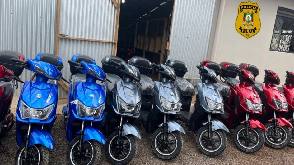 Esta &eacute; uma descri&ccedil;&atilde;o detalhada da imagem para fins de acessibilidade:

Vis&atilde;o Geral
A imagem apresenta uma fileira de motocicletas el&eacute;tricas ou scooters novas, estacionadas em frente a uma instala&ccedil;&atilde;o da Pol&iacute;cia Penal do Rio Grande do Sul.

As Motocicletas
Cores e Modelos: Est&atilde;o alinhadas cerca de oito motocicletas em tr&ecirc;s cores distintas: azul vibrante &agrave; esquerda, cinza met&aacute;lico ao centro e vermelho &agrave; direita.

Detalhes: Todas possuem um ba&uacute; de carga preto (top case) instalado na parte traseira. Algumas ainda mant&ecirc;m o pl&aacute;stico de prote&ccedil;&atilde;o nos assentos, indicando serem ve&iacute;culos novos.

O Ambiente
Edifica&ccedil;&atilde;o: As motos est&atilde;o estacionadas em um p&aacute;tio de brita em frente a um galp&atilde;o com paredes de metal galvanizado e uma porta de correr parcialmente aberta.

Identifica&ccedil;&atilde;o Oficial: Na parede lateral clara, &agrave; direita, destaca-se o bras&atilde;o oficial amarelo e verde da Pol&iacute;cia Penal RS.

Contexto: A cena sugere a moderniza&ccedil;&atilde;o da frota ou a implementa&ccedil;&atilde;o de novos recursos de mobilidade para o sistema prisional, em conformidade com as normas mencionadas no t&iacute;tulo do arquivo.