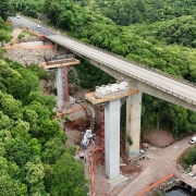 Esta é uma descrição detalhada da imagem para fins de acessibilidade:

Visão Geral
A imagem apresenta uma vista aérea de grande ângulo (provavelmente capturada por um drone) de uma importante obra de infraestrutura rodoviária. O cenário mostra a construção de novos pilares para uma ponte em meio a um vale de vegetação densa e preservada.

A Ponte e a Obra
Ponte Existente: Uma longa ponte de concreto cinza atravessa o vale diagonalmente, da esquerda para a direita. Veículos leves podem ser vistos circulando sobre ela.

Novas Estruturas: Paralelamente à ponte atual, estão sendo erguidos novos pilares de sustentação em concreto.

Atividade de Construção:

No topo dos novos pilares, há plataformas de trabalho cercadas por andaimes e formas de madeira. É possível avistar operários em uniformes laranjas trabalhando nessas alturas.

Na base da estrutura central, está posicionado um caminhão bomba de concreto branco com braços estendidos.

Estruturas metálicas amarelas (provavelmente treliças de lançamento ou guindastes) estão em uso para o posicionamento de vigas na extremidade inferior direita da obra.

Preparação do Terreno: Áreas de encosta próximas à cabeceira da ponte exibem terraplanagem e contenções feitas com telas de proteção laranjas.

O Entorno Natural
Vegetação: O vale é coberto por uma floresta verde exuberante e contínua, que contrasta com o tom cinza das estruturas de concreto.

Geografia: O terreno é acidentado, com morros íngremes. Um caminho de terra serpenteia o fundo do vale, próximo ao leito do arroio.