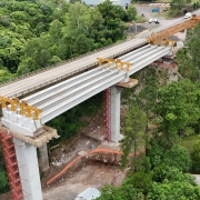 Esta é uma descrição detalhada da imagem para fins de acessibilidade:

Visão Geral
A imagem apresenta uma vista aérea de grande ângulo (provavelmente capturada por um drone) de uma importante obra de infraestrutura rodoviária. O cenário mostra a construção de novos pilares para uma ponte em meio a um vale de vegetação densa e preservada.

A Ponte e a Obra
Ponte Existente: Uma longa ponte de concreto cinza atravessa o vale diagonalmente, da esquerda para a direita. Veículos leves podem ser vistos circulando sobre ela.

Novas Estruturas: Paralelamente à ponte atual, estão sendo erguidos novos pilares de sustentação em concreto.

Atividade de Construção:

No topo dos novos pilares, há plataformas de trabalho cercadas por andaimes e formas de madeira. É possível avistar operários em uniformes laranjas trabalhando nessas alturas.

Na base da estrutura central, está posicionado um caminhão bomba de concreto branco com braços estendidos.

Estruturas metálicas amarelas (provavelmente treliças de lançamento ou guindastes) estão em uso para o posicionamento de vigas na extremidade inferior direita da obra.

Preparação do Terreno: Áreas de encosta próximas à cabeceira da ponte exibem terraplanagem e contenções feitas com telas de proteção laranjas.

O Entorno Natural
Vegetação: O vale é coberto por uma floresta verde exuberante e contínua, que contrasta com o tom cinza das estruturas de concreto.

Geografia: O terreno é acidentado, com morros íngremes. Um caminho de terra serpenteia o fundo do vale, próximo ao leito do arroio.