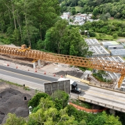 Esta é uma descrição detalhada da imagem para fins de acessibilidade:

Visão Geral
A imagem apresenta uma vista aérea de grande ângulo (provavelmente capturada por um drone) de uma importante obra de infraestrutura rodoviária. O cenário mostra a construção de novos pilares para uma ponte em meio a um vale de vegetação densa e preservada.

A Ponte e a Obra
Ponte Existente: Uma longa ponte de concreto cinza atravessa o vale diagonalmente, da esquerda para a direita. Veículos leves podem ser vistos circulando sobre ela.

Novas Estruturas: Paralelamente à ponte atual, estão sendo erguidos novos pilares de sustentação em concreto.

Atividade de Construção:

No topo dos novos pilares, há plataformas de trabalho cercadas por andaimes e formas de madeira. É possível avistar operários em uniformes laranjas trabalhando nessas alturas.

Na base da estrutura central, está posicionado um caminhão bomba de concreto branco com braços estendidos.

Estruturas metálicas amarelas (provavelmente treliças de lançamento ou guindastes) estão em uso para o posicionamento de vigas na extremidade inferior direita da obra.

Preparação do Terreno: Áreas de encosta próximas à cabeceira da ponte exibem terraplanagem e contenções feitas com telas de proteção laranjas.

O Entorno Natural
Vegetação: O vale é coberto por uma floresta verde exuberante e contínua, que contrasta com o tom cinza das estruturas de concreto.

Geografia: O terreno é acidentado, com morros íngremes. Um caminho de terra serpenteia o fundo do vale, próximo ao leito do arroio.