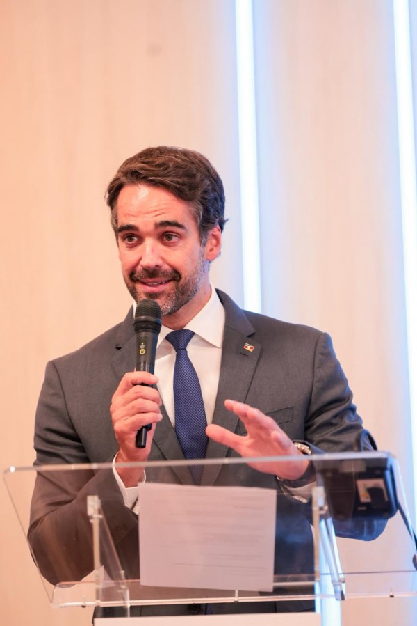 Esta é uma descrição detalhada da imagem para fins de acessibilidade:

Visão Geral
A imagem é um retrato em plano médio (do peito para cima) do governador Eduardo Leite, capturado durante um pronunciamento oficial em um ambiente interno.

O Governador e sua Ação
Aparência e Traje: O governador, um homem de pele clara com cabelos e barba curta castanhos, veste um traje formal composto por terno cinza escuro, camisa branca e gravata azul texturizada. Na lapela esquerda do paletó, há um pequeno botom com as cores da bandeira do Rio Grande do Sul.

Gesto e Expressão: Ele segura um microfone preto com a mão direita próximo ao rosto, enquanto a mão esquerda está erguida em um gesto de ênfase. Sua expressão é atenta e comunicativa, com o olhar direcionado para o lado.

O Ambiente e Composição
Púlpito: À frente do governador, vê-se a parte superior de um púlpito de acrílico transparente, onde estão apoiadas folhas de papel com anotações para o discurso.

Plano de Fundo: O fundo é composto por painéis de madeira clara com acabamento minimalista, destacados por faixas verticais de luz branca brilhante, o que confere um aspecto moderno e sóbrio à cena.