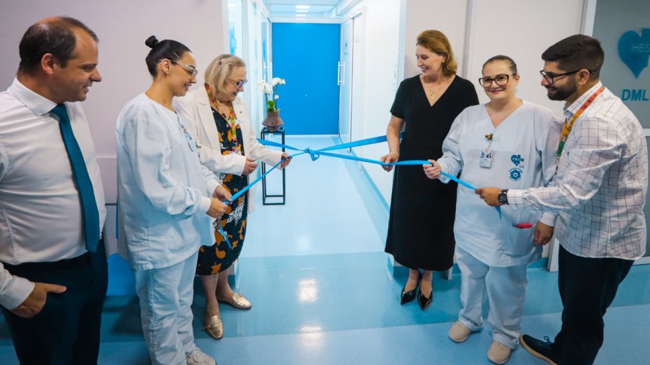Foto posada em ambiente hospitalar de paredes brancas e piso azul. Quatro mulheres e um homem est&atilde;o de p&eacute; e seguram uma fita azul amarrada, que ir&atilde;o descerrar. Um homem assiste &agrave; cena.
