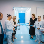 Foto posada em ambiente hospitalar de paredes brancas e piso azul. Quatro mulheres e um homem est&atilde;o de p&eacute; e seguram uma fita azul amarrada, que ir&atilde;o descerrar. Um homem assiste &agrave; cena.