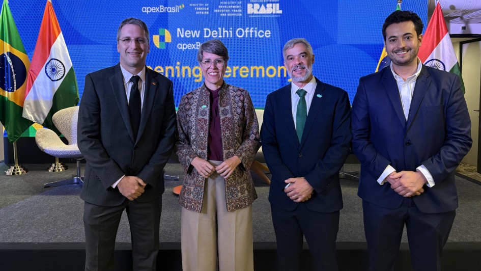 Vis&atilde;o Geral
A imagem mostra quatro pessoas (tr&ecirc;s homens e uma mulher) posando para uma foto oficial sobre um palco durante uma cerim&ocirc;nia de inaugura&ccedil;&atilde;o. Eles est&atilde;o posicionados &agrave; frente de um grande tel&atilde;o azul vibrante que identifica o evento.

As Pessoas (da esquerda para a direita)
Primeiro Homem: Veste terno cinza escuro, camisa branca, gravata preta e usa &oacute;culos de grau.

Mulher: No centro-esquerda, possui cabelos curtos grisalhos e usa &oacute;culos. Veste uma jaqueta de tecido estruturado com estampas detalhadas em tons de marrom e bege sobre uma blusa roxa, combinada com cal&ccedil;a bege de corte largo.

Segundo Homem: No centro-direita, possui barba grisalha e veste terno azul-marinho com uma gravata verde.

Terceiro Homem: Na extremidade direita, &eacute; mais jovem e veste um terno azul-marinho com camisa branca, sem gravata.

O Cen&aacute;rio e Identifica&ccedil;&atilde;o
Tel&atilde;o de Fundo: De cor azul brilhante com textura de pequenos pontos, exibe os textos em branco e amarelo: "New Delhi Office", "apexBrasil" e "opening ceremony". Na parte superior, constam logotipos da ApexBrasil, do Minist&eacute;rio das Rela&ccedil;&otilde;es Exteriores e do Governo Federal do Brasil.

Bandeiras: Pares das bandeiras do Brasil e da &Iacute;ndia est&atilde;o posicionados em mastros nas duas extremidades do palco.

Palco: O ch&atilde;o &eacute; revestido por um carpete cinza escuro, e ao fundo, atr&aacute;s das autoridades, veem-se duas poltronas brancas de design moderno (estilo "Swan").